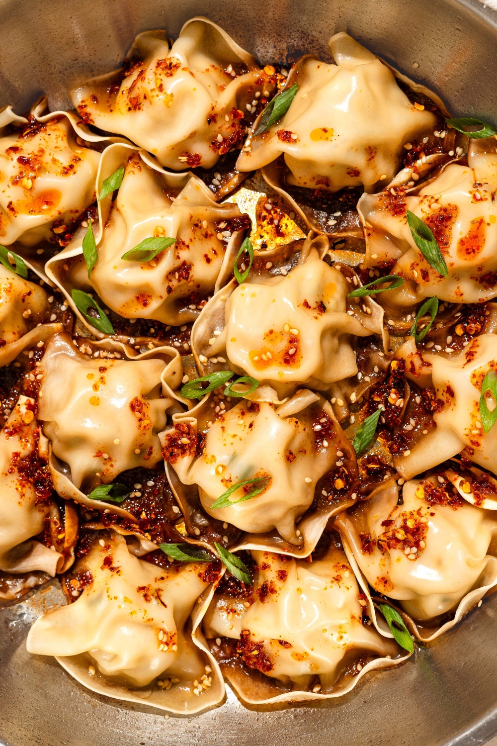 A skillet with blanket dumplings topped with chili crisp, sesame seeds, and sliced green onion. The skillet is on a tan counter.