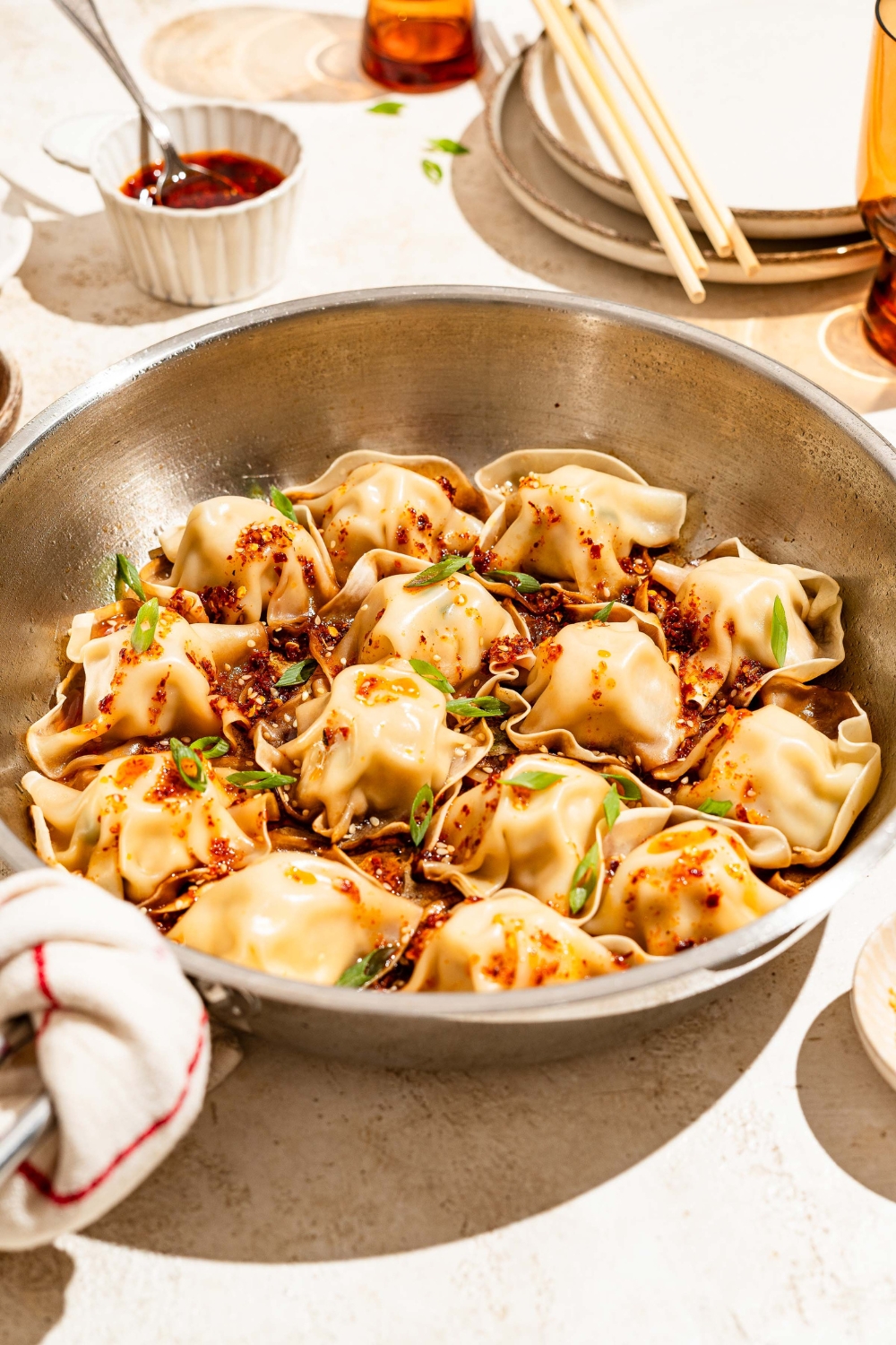 A skillet with blanket dumplings topped with chili crisp, sesame seeds, and sliced green onion. The skillet is on a tan counter.