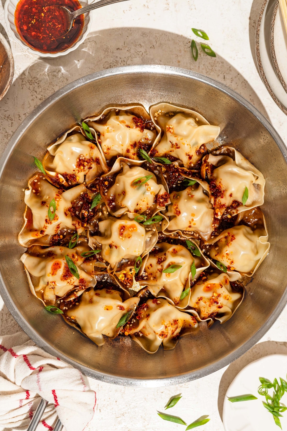 A skillet with blanket dumplings topped with chili crisp, sesame seeds, and sliced green onion. The skillet is on a tan counter with a bowl of chili crisp.