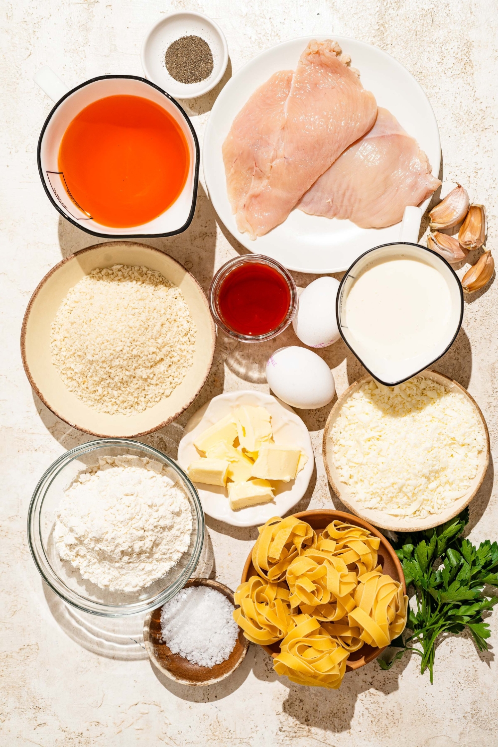 A tan counter with several bowls containing ingredients to make Buffalo chicken alfredo including hot sauce, chicken, butter, fettuccine pasta, heavy cream, parmesan cheese, flour, egg, breadcrumbs, garlic, and more.