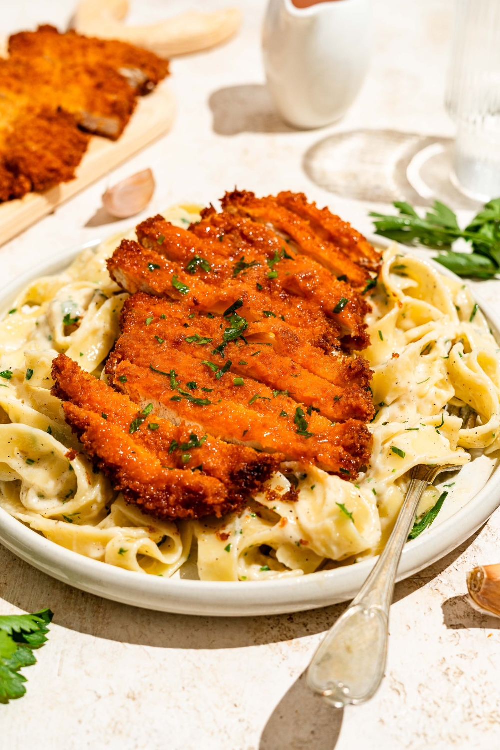 A white plate with crispy Buffalo chicken sliced and served over fettuccine alfredo. The plate is served with a fork and garnished with fresh parsley. The plate is on a tan counter with a board of crispy Buffalo chicken.