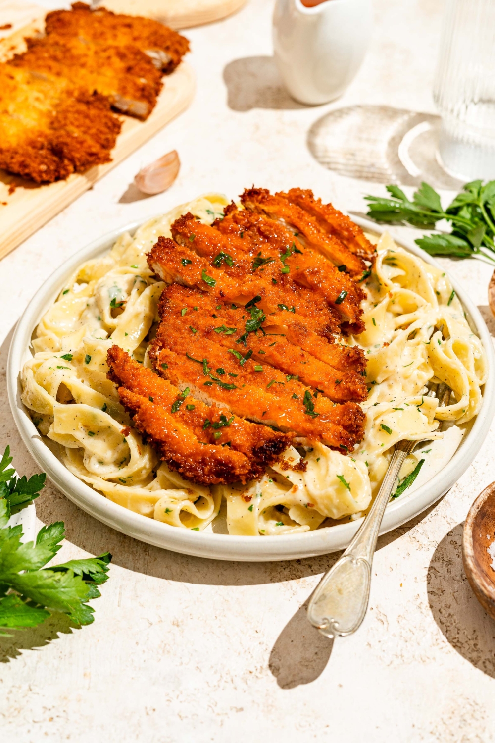A white plate with crispy Buffalo chicken sliced and served over fettuccine alfredo. The plate is served with a fork and garnished with fresh parsley. The plate is on a tan counter with a board of crispy Buffalo chicken.