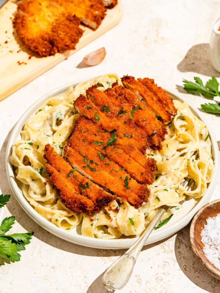 A white plate with crispy Buffalo chicken sliced and served over fettuccine alfredo. The plate is served with a fork and garnished with fresh parsley. The plate is on a tan counter with a board of crispy Buffalo chicken.