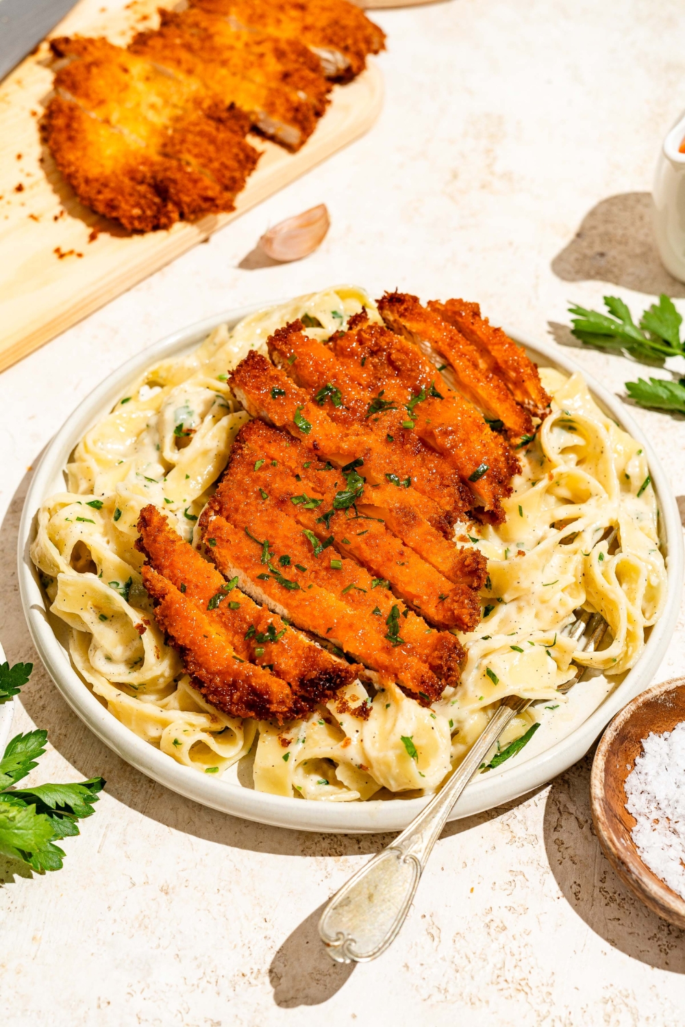 A white plate with crispy Buffalo chicken sliced and served over fettuccine alfredo. The plate is served with a fork and garnished with fresh parsley. The plate is on a tan counter with a board of crispy Buffalo chicken.