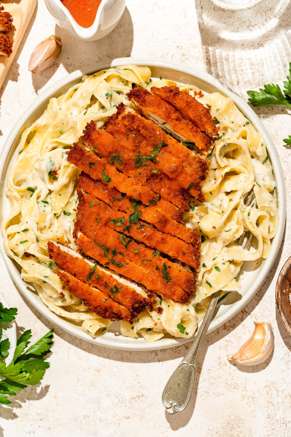 A white plate with crispy Buffalo chicken sliced and served over fettuccine alfredo. The plate is served with a fork and garnished with fresh parsley. The plate is on a tan counter with a board of crispy Buffalo chicken.