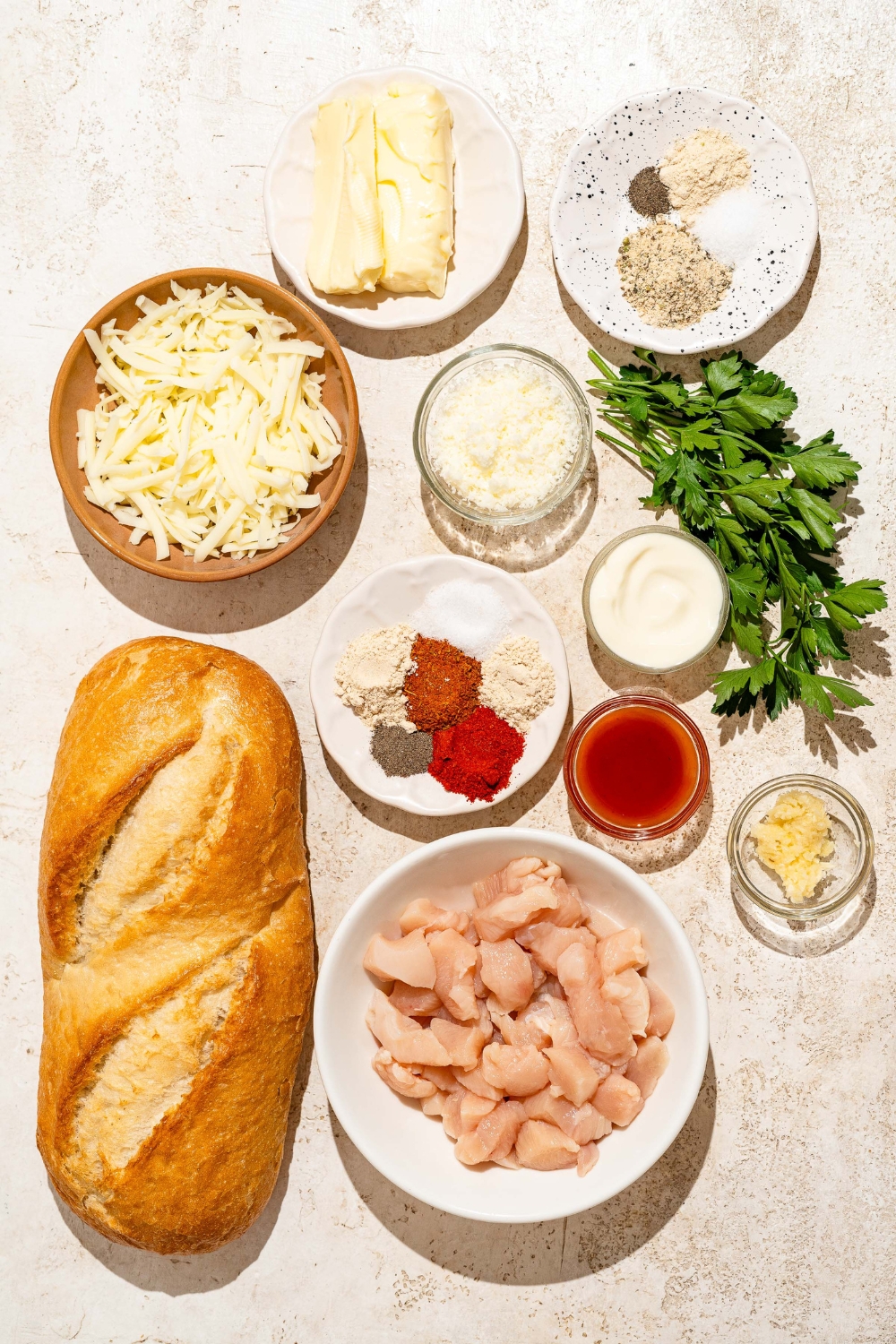 A tan counter with ingredients to make Buffalo chicken garlic bread including Italian bread, chicken, mozzarella cheese, garlic, butter, sour cream, Buffalo sauce, and seasonings.