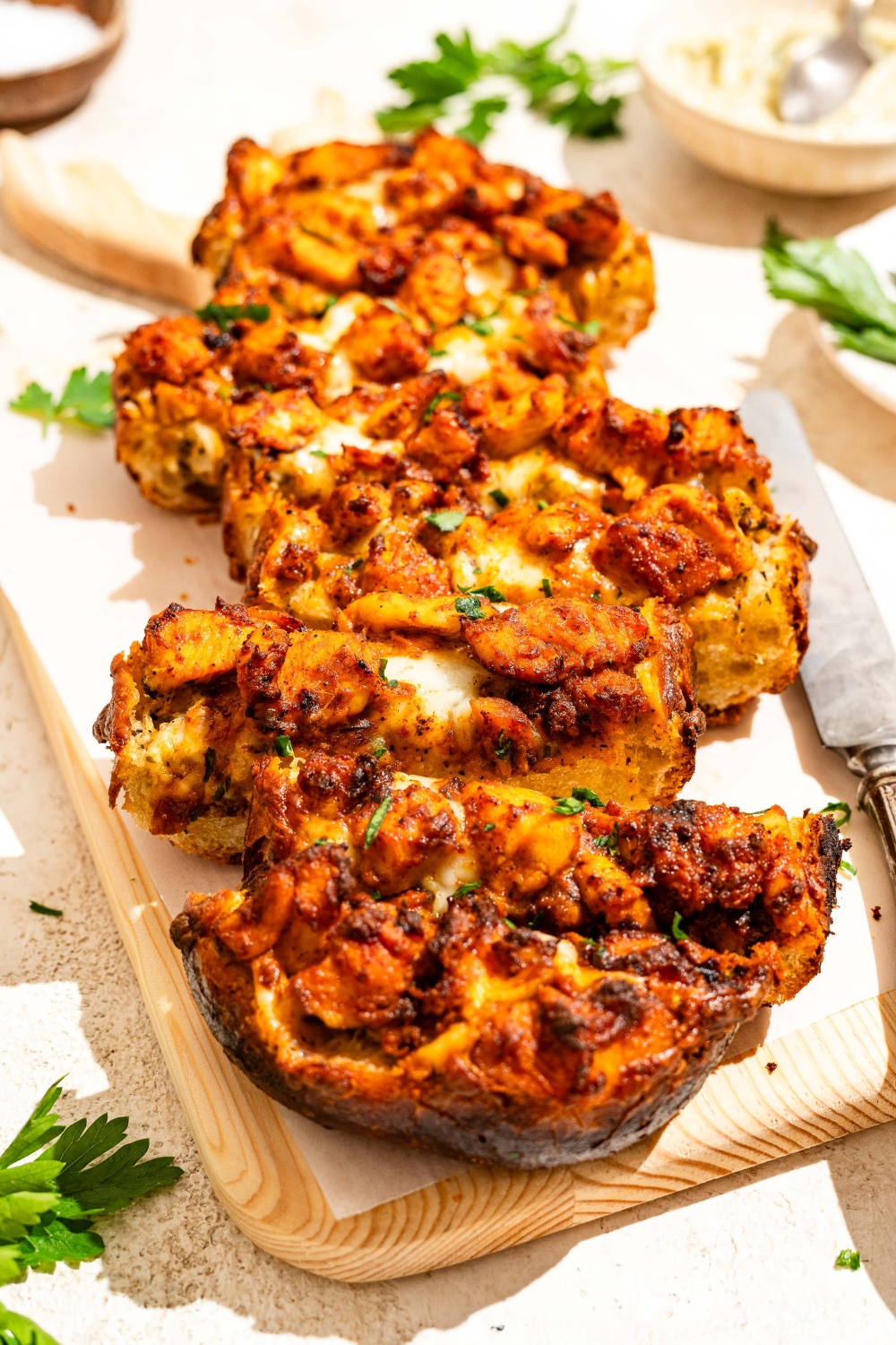 A wooden board lined with parchment paper with Buffalo chicken garlic bread garnished with fresh parsley and sliced in strips. The board is on a tan counter.