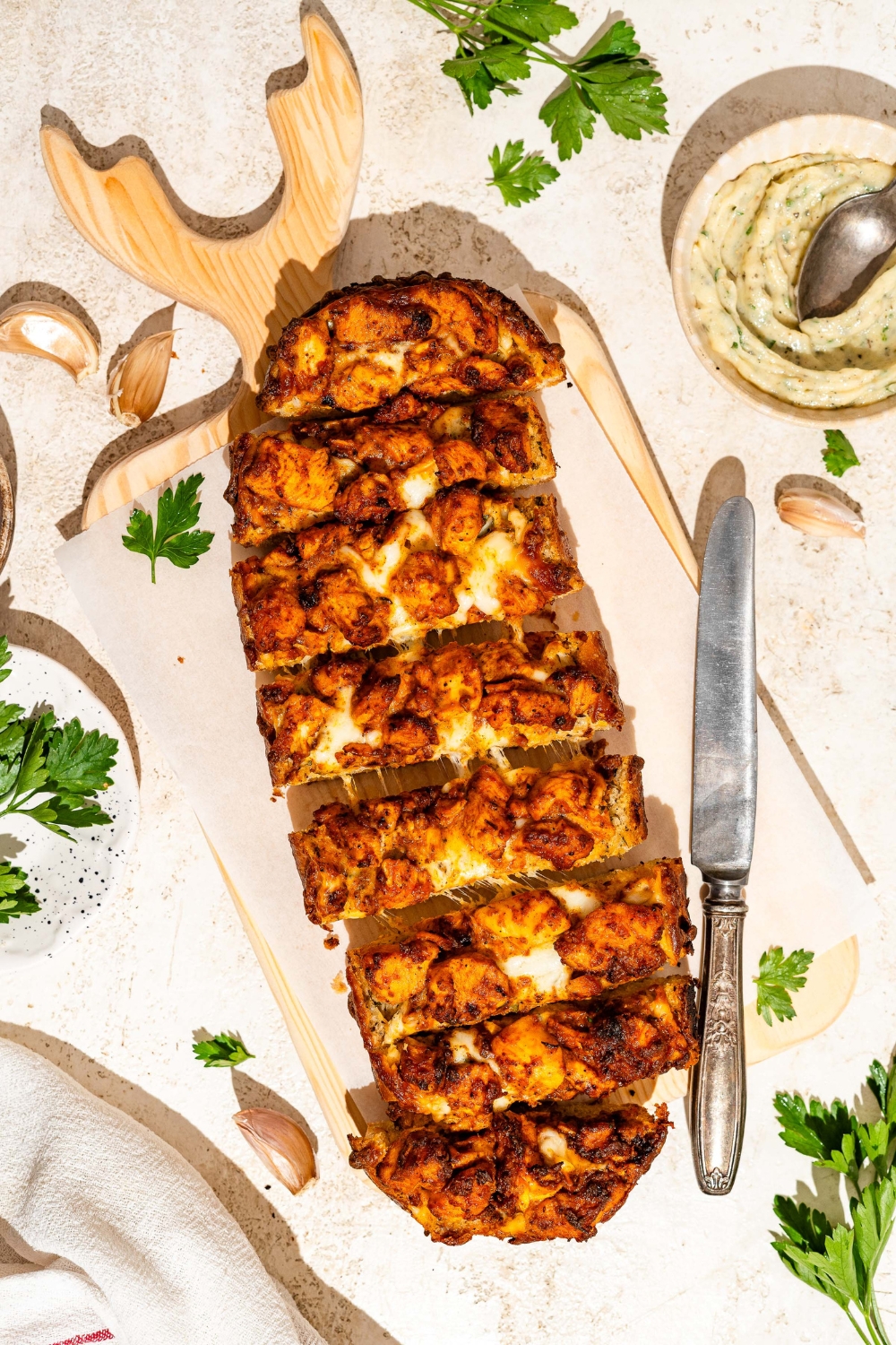 A wooden board lined with parchment paper with Buffalo chicken garlic bread garnished with fresh parsley and sliced in strips. The board is on a tan counter with a knife and bowl of garlic butter.