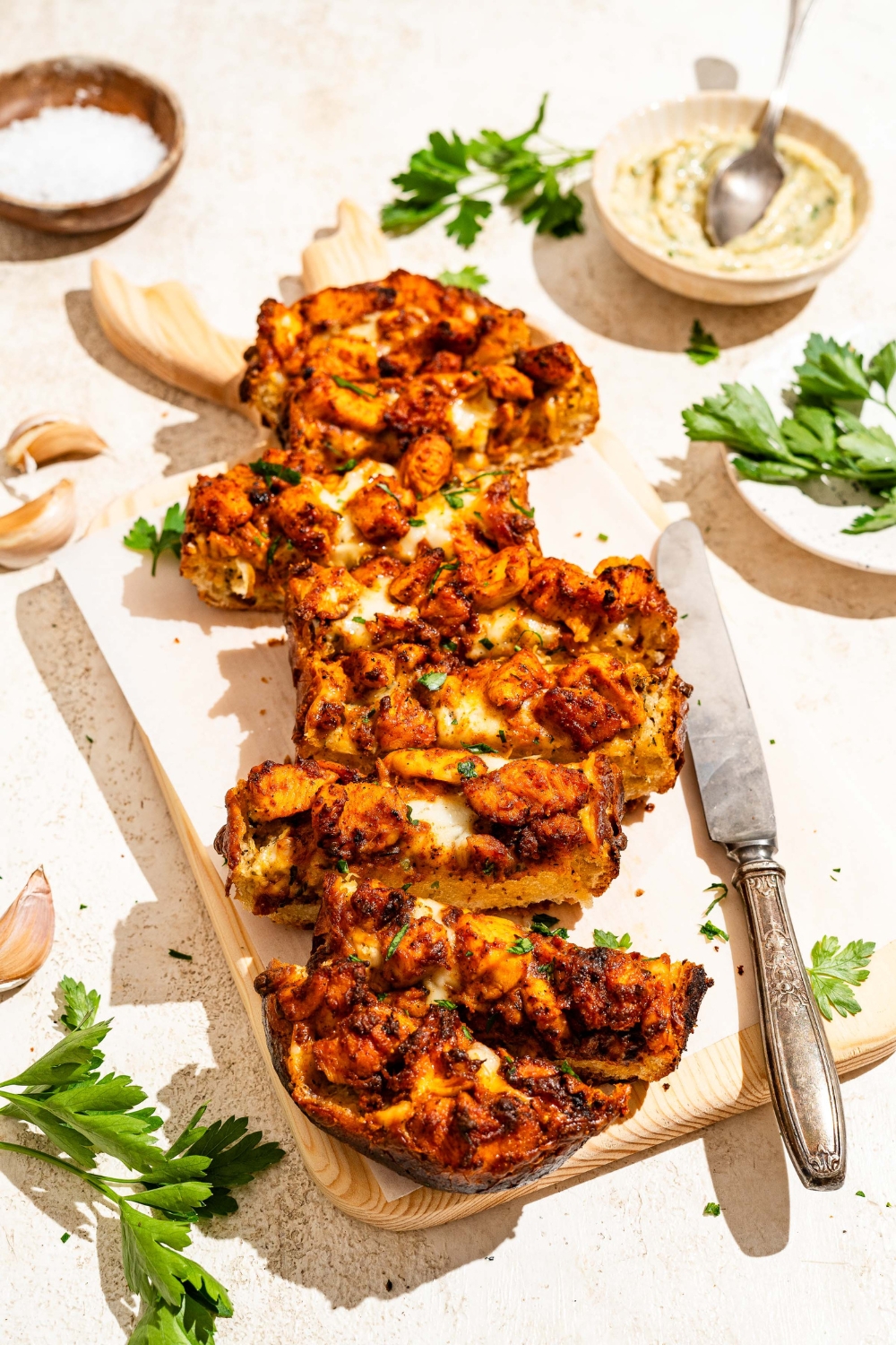 A wooden board lined with parchment paper with Buffalo chicken garlic bread garnished with fresh parsley and sliced in strips. The board is on a tan counter with a knife and bowl of garlic butter.