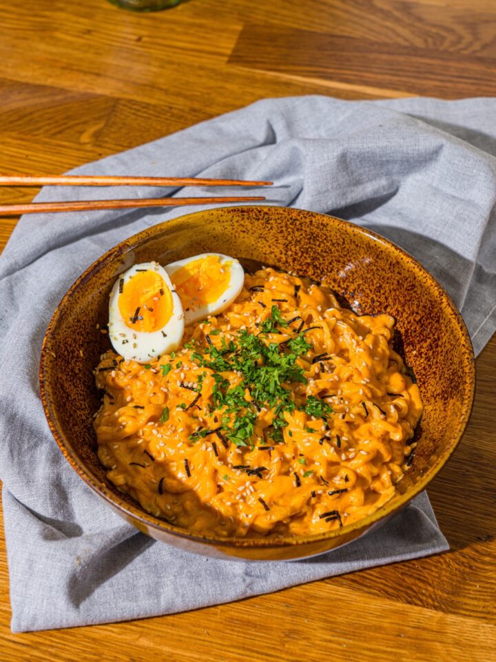 A bowl of Buldak ramen with soft boiled egg and garnished with fresh parsley and sesame seeds. The bowl is on a wooden counter with a blue cloth napkin and a pair of chopsticks.