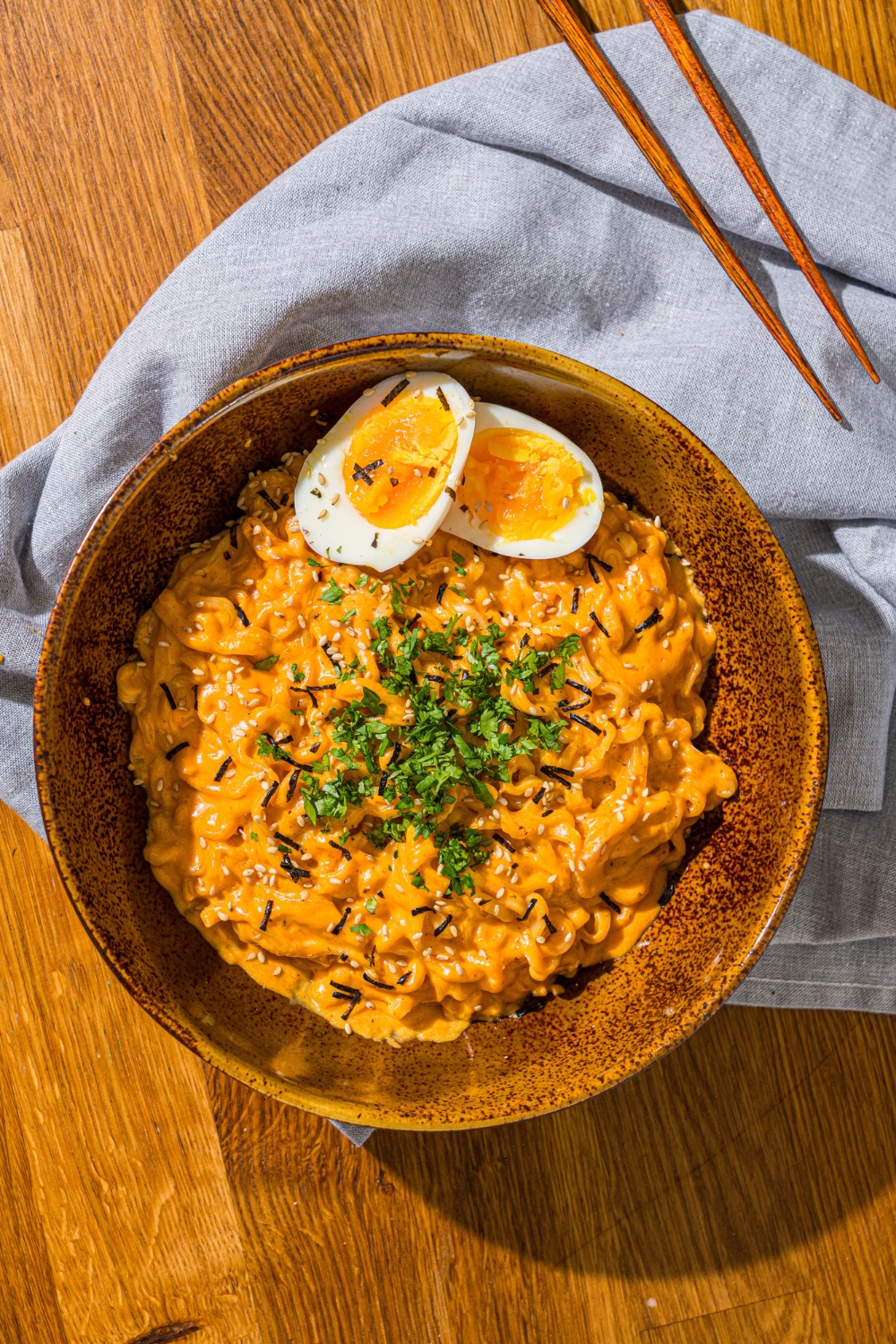 A bowl of Buldak ramen with soft boiled egg and garnished with fresh parsley and sesame seeds. The bowl is on a wooden counter with a blue cloth napkin and a pair of chopsticks.