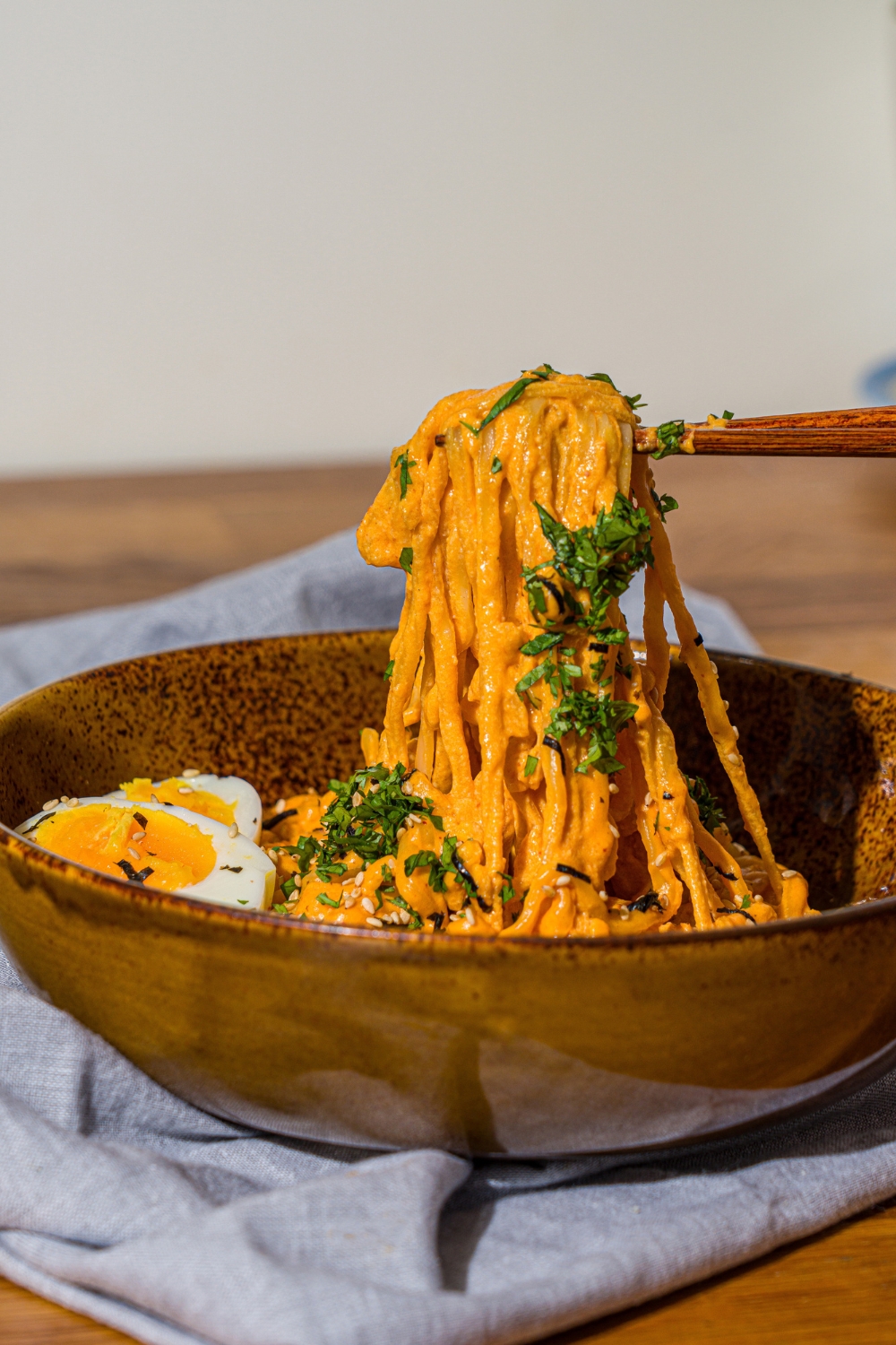 A bowl of Buldak ramen with soft boiled egg and garnished with fresh parsley and sesame seeds. A pair of chopsticks are pulling noodles from the bowl. The bowl is on a wooden counter with a blue cloth napkin.