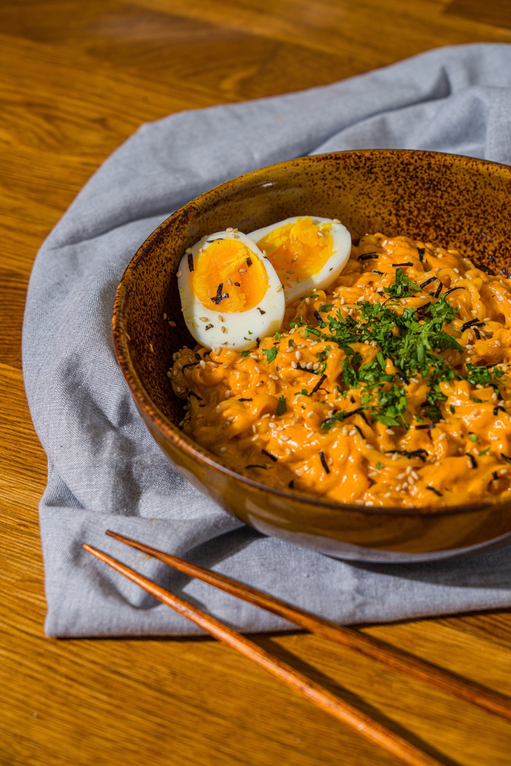 A bowl of Buldak ramen with soft boiled egg and garnished with fresh parsley and sesame seeds. The bowl is on a wooden counter with a blue cloth napkin and a pair of chopsticks.