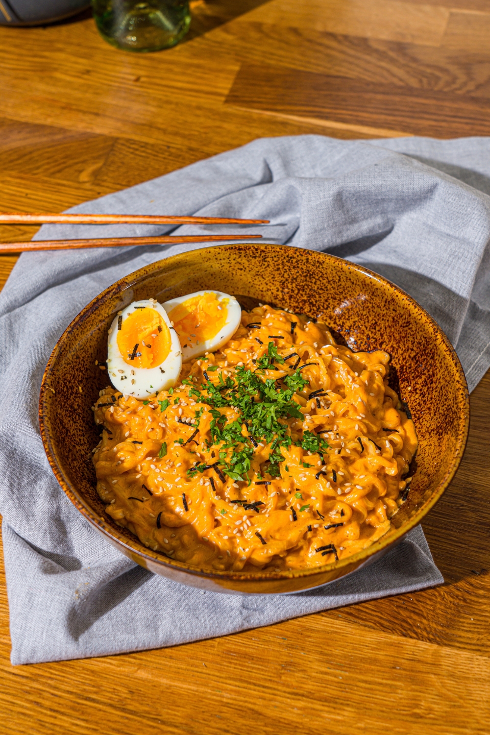 A bowl of Buldak ramen with soft boiled egg and garnished with fresh parsley and sesame seeds. The bowl is on a wooden counter with a blue cloth napkin and a pair of chopsticks.