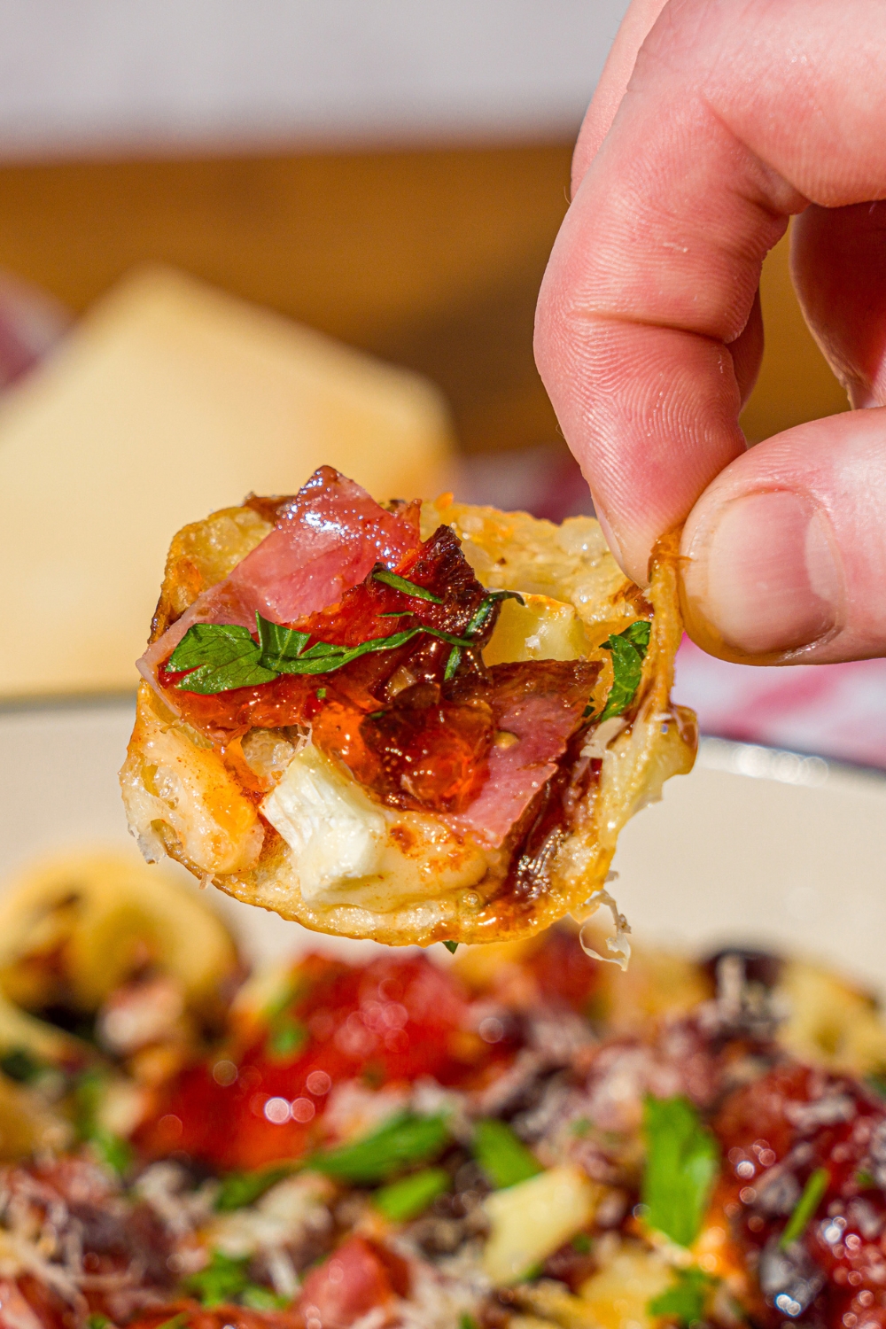 A close up of a hand holding a charcuterie nacho topped with balsamic glaze and fresh bail. There is a dish of nachos blurred in the background.