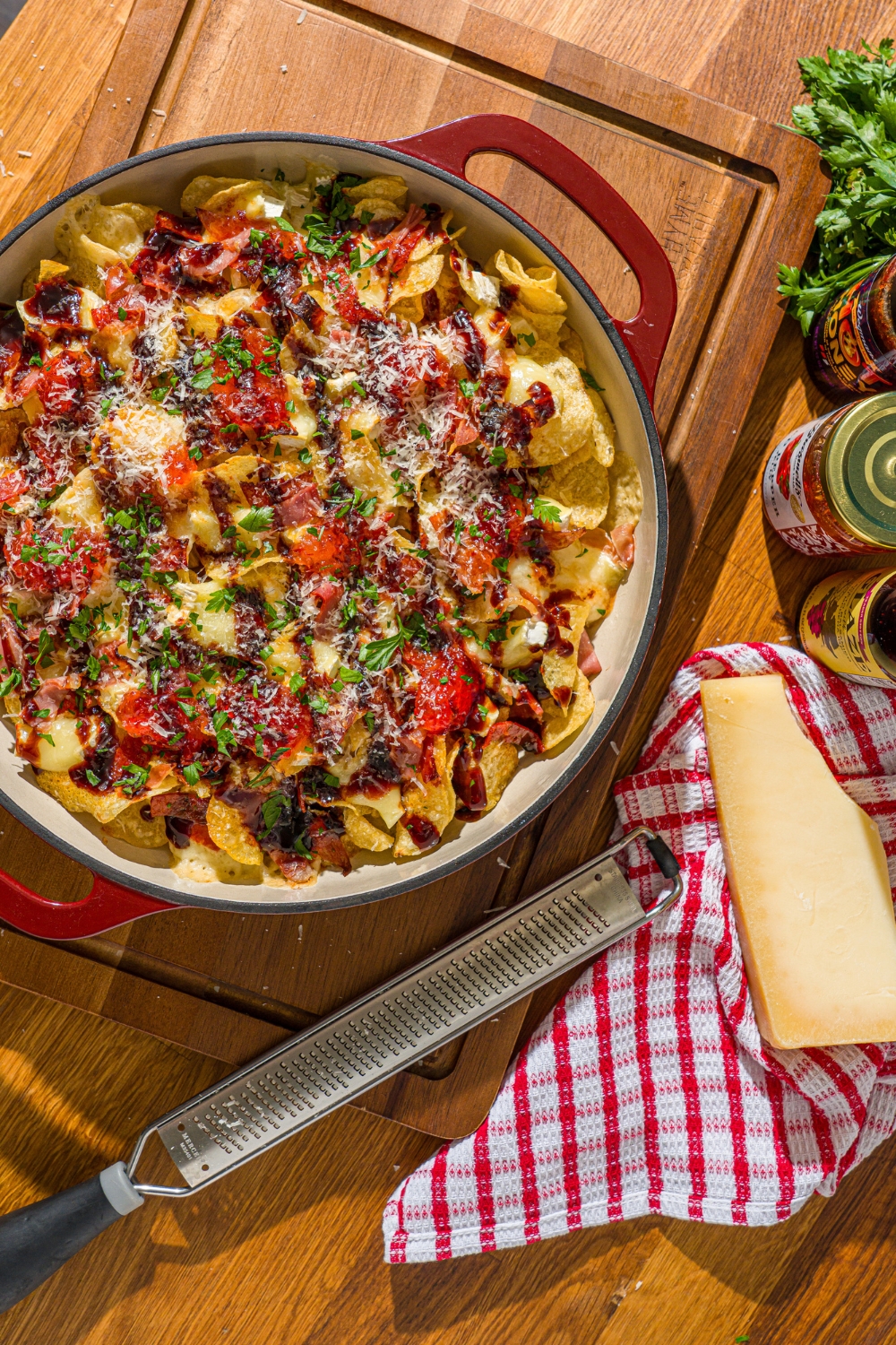 A baking dish with baked charcuterie nachos topped with balsamic glaze, shredded cheese, and fresh basil. The dish is on a wooden cutting board on a wooden counter with a red striped napkin.