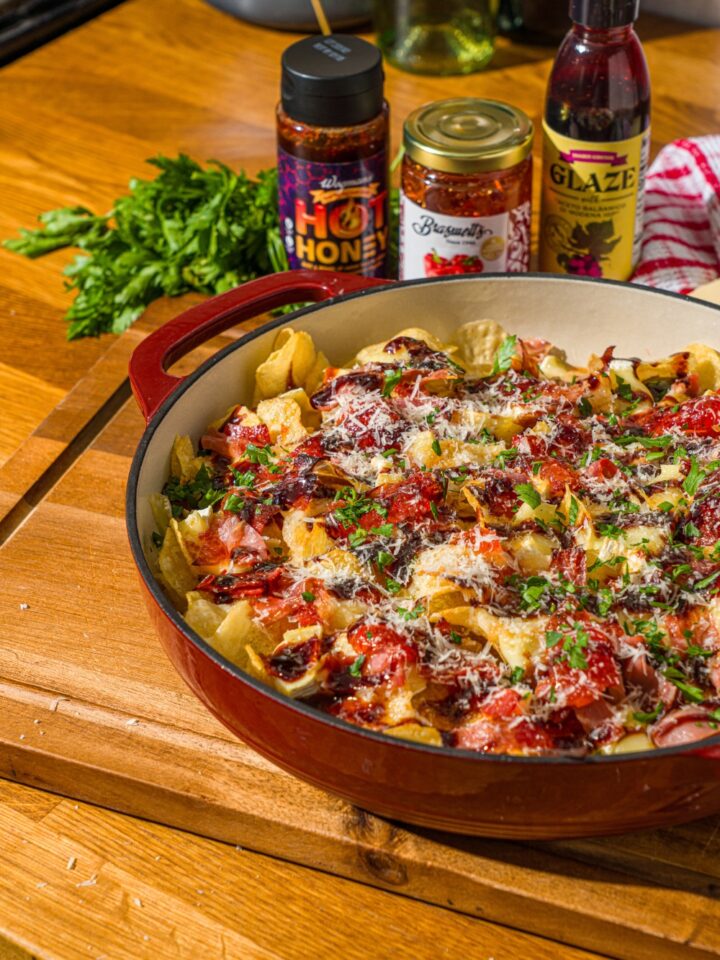 A baking dish with baked charcuterie nachos topped with balsamic glaze, shredded cheese, and fresh basil. A hand has taken a chip from the dish. The dish is on a wooden counter with jars of toppings.