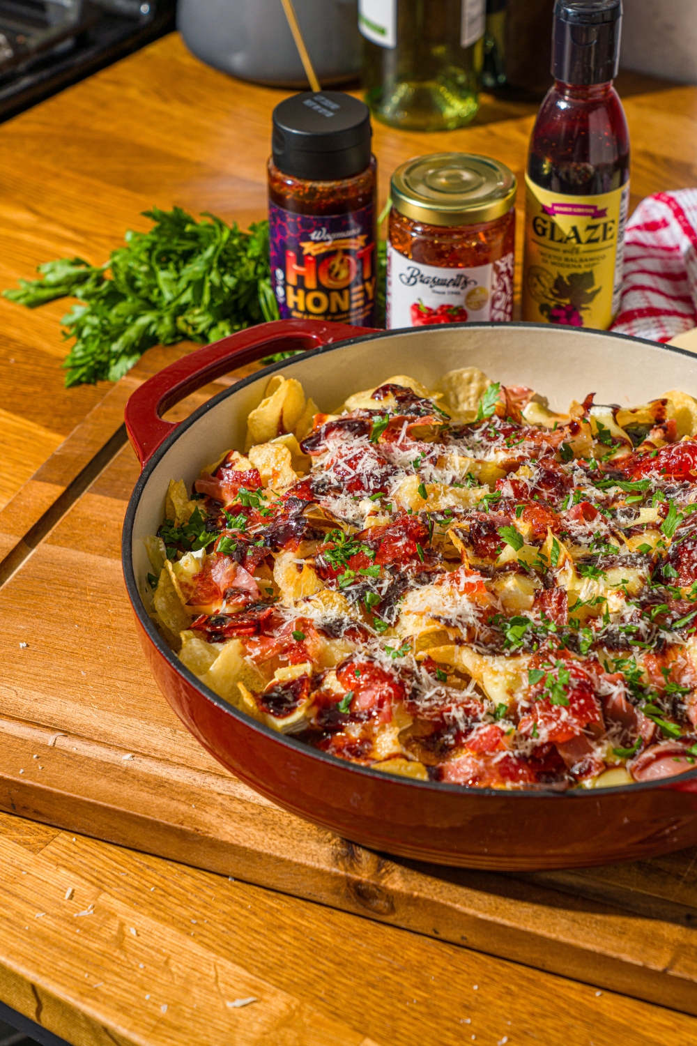 A baking dish with baked charcuterie nachos topped with balsamic glaze, shredded cheese, and fresh basil. A hand has taken a chip from the dish. The dish is on a wooden counter with jars of toppings.