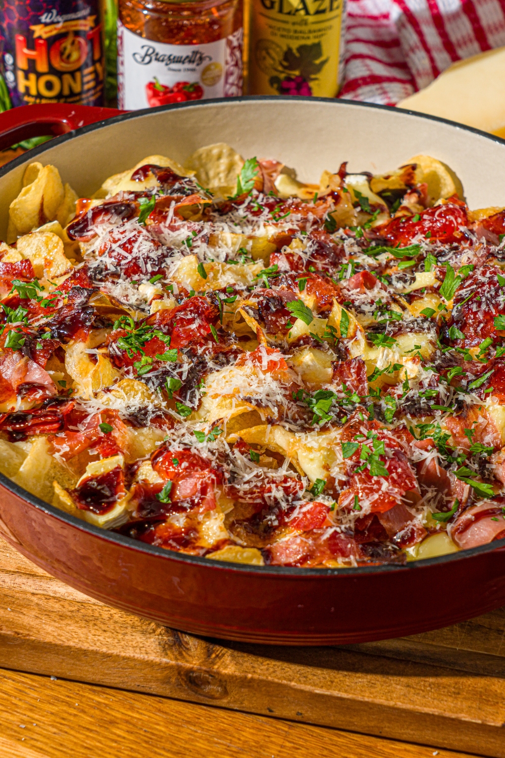 A baking dish with baked charcuterie nachos topped with balsamic glaze, shredded cheese, and fresh basil. The dish is on a wooden counter with a red striped napkin.