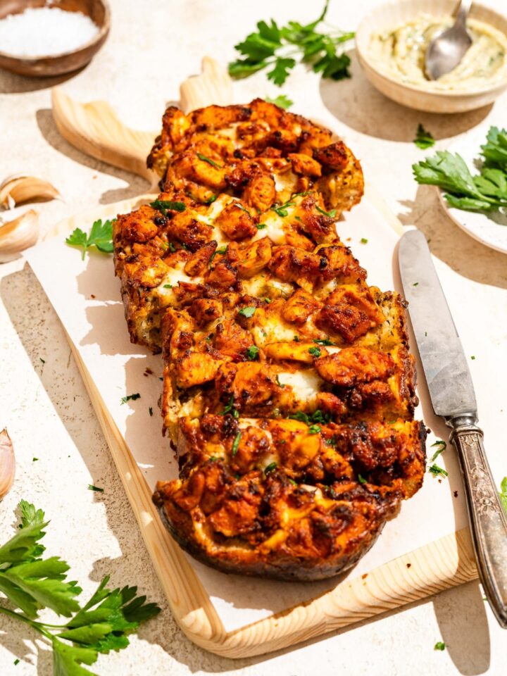 A wooden board lined with parchment paper with Buffalo chicken garlic bread garnished with fresh parsley and sliced in strips. The board is on a tan counter with a knife and a bowl of garlic butter.