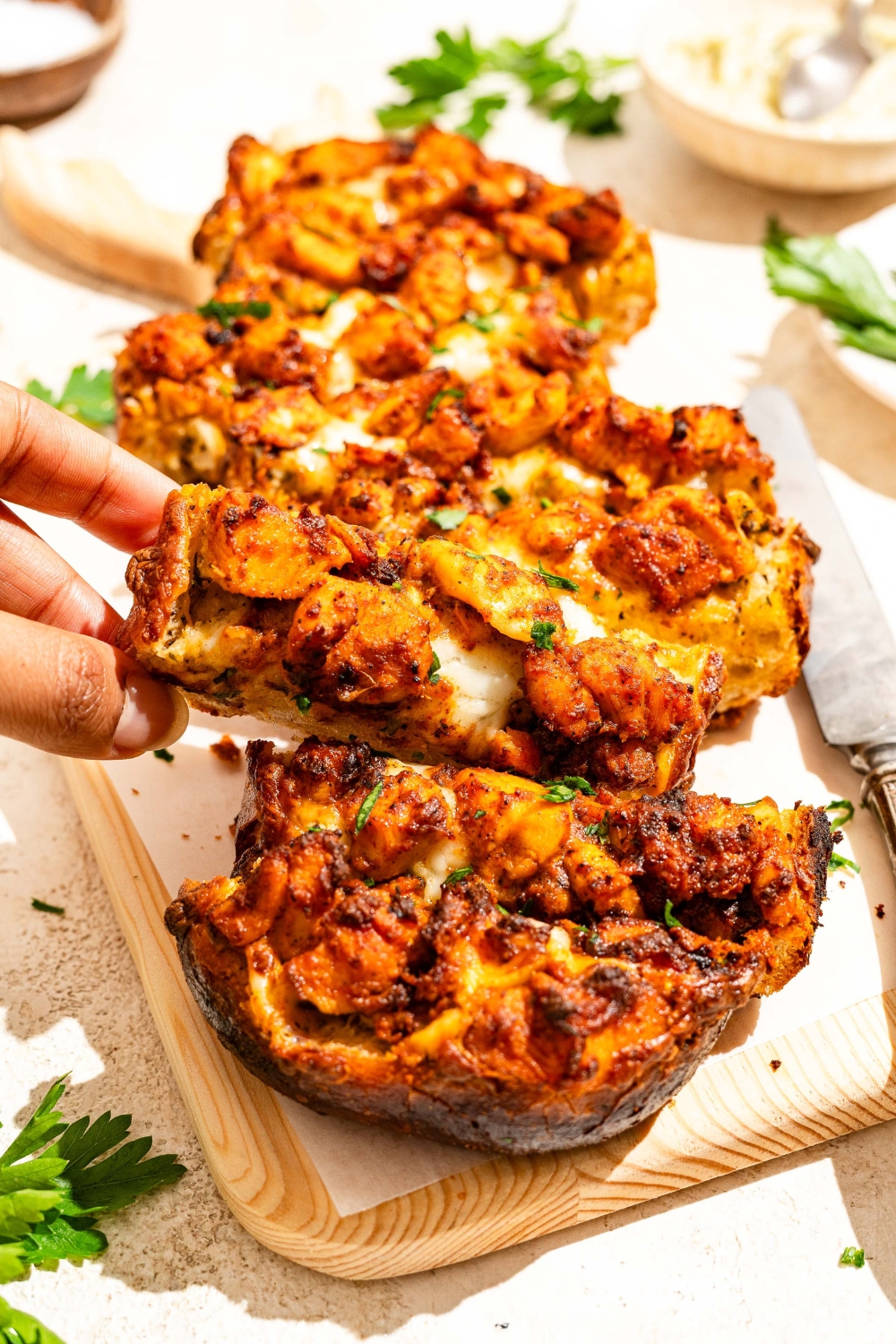 A wooden board lined with parchment paper with Buffalo chicken garlic bread garnished with fresh parsley and sliced in strips. A hand is taking a slice of garlic bread. The board is on a tan counter with a knife.