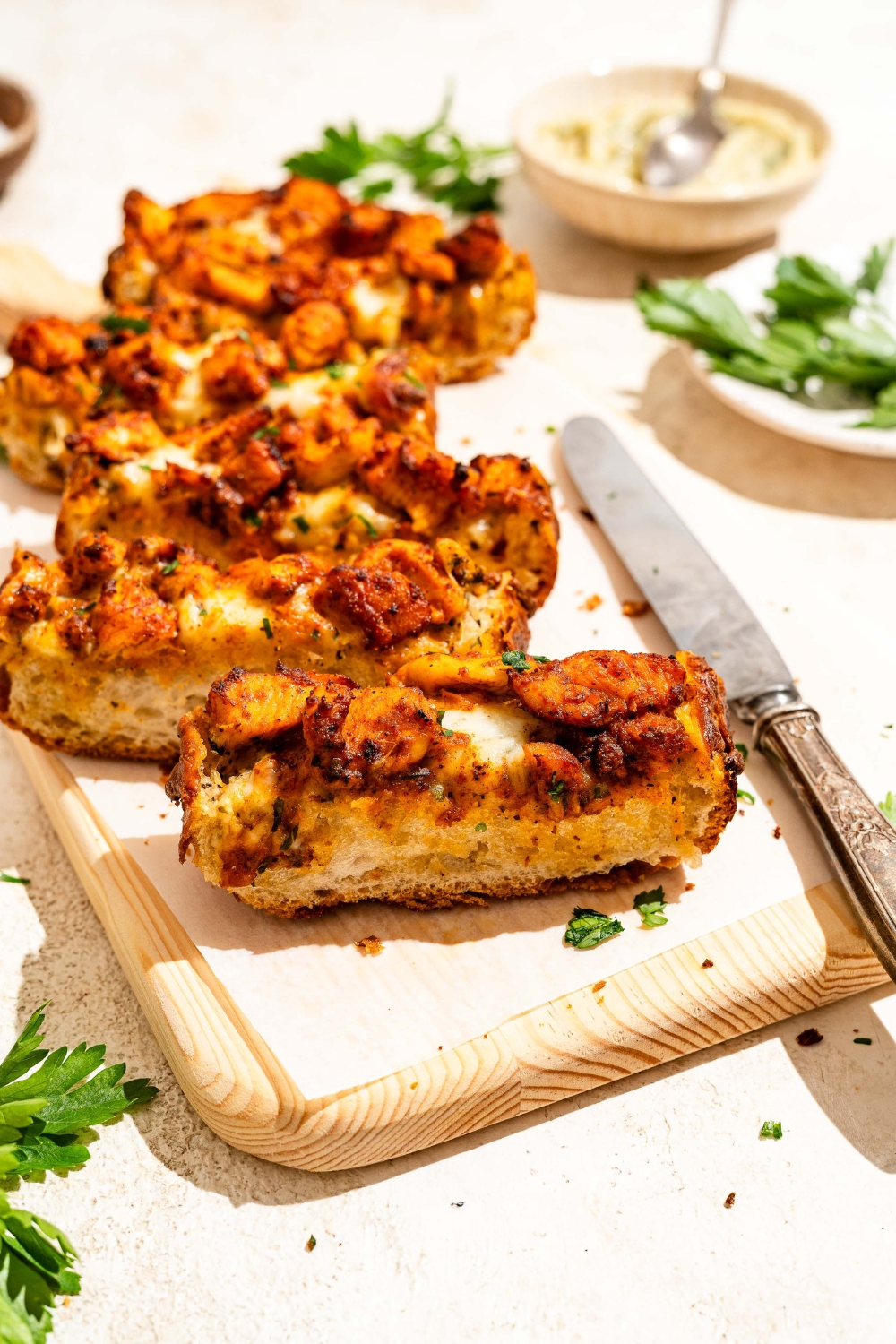 A wooden board lined with parchment paper with Buffalo chicken garlic bread garnished with fresh parsley and sliced in strips. The board is on a tan counter with a knife and a bowl of garlic butter.