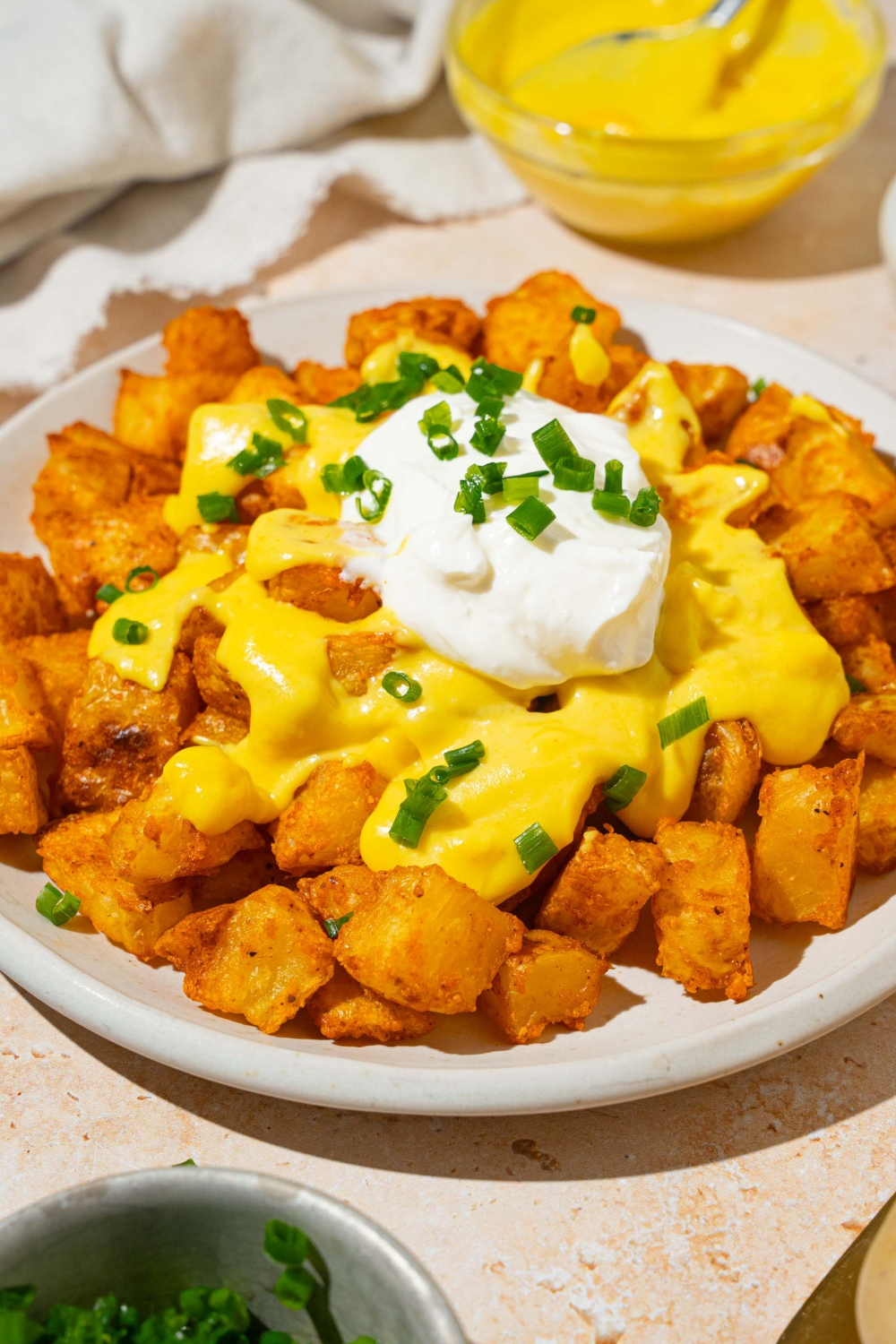 A white plate with copycat cheesy fiesta potatoes topped with taco bell nacho cheese sauce, sour cream, and sliced green onions. The plate is on a tan counter with a bowl of nacho cheese sauce and white cloth napkin.