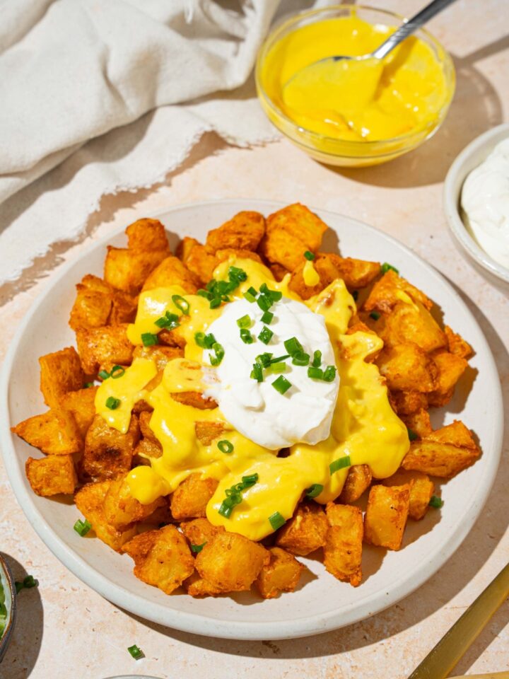 A white plate with copycat cheesy fiesta potatoes topped with taco bell nacho cheese sauce, sour cream, and sliced green onions. The plate is on a tan counter with a bowl of nacho cheese sauce and white cloth napkin.