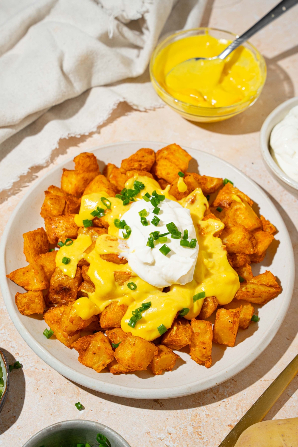 A white plate with copycat cheesy fiesta potatoes topped with taco bell nacho cheese sauce, sour cream, and sliced green onions. The plate is on a tan counter with a bowl of nacho cheese sauce and white cloth napkin.