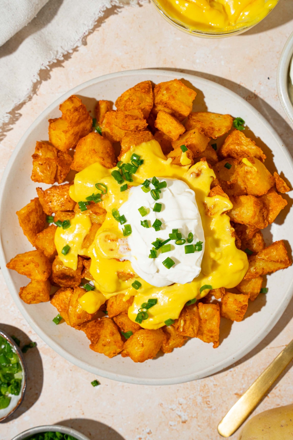 A white plate with copycat cheesy fiesta potatoes topped with taco bell nacho cheese sauce, sour cream, and sliced green onions. The plate is on a tan counter with a bowl of nacho cheese sauce and white cloth napkin.
