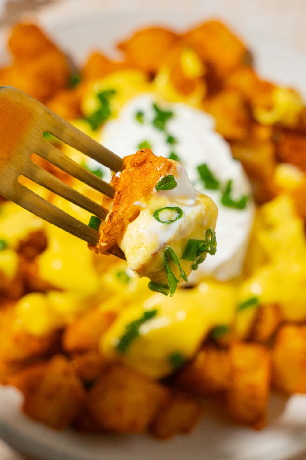 A close up of a fork with a bite of cheesy fiesta potatoes garnished with sliced green onions. There is a plate of potatoes topped with taco bell nacho cheese and sour cream blurred in the background.