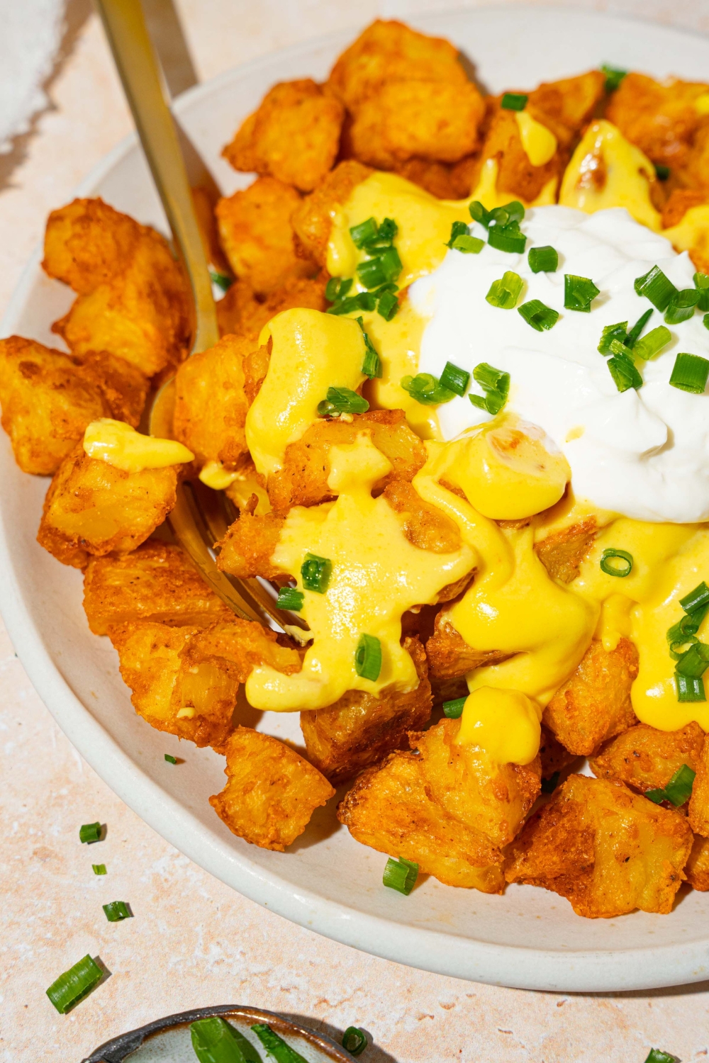 A white plate with copycat cheesy fiesta potatoes topped with taco bell nacho cheese sauce, sour cream, and sliced green onions. There is a fork in the potatoes. The plate is on a tan counter.