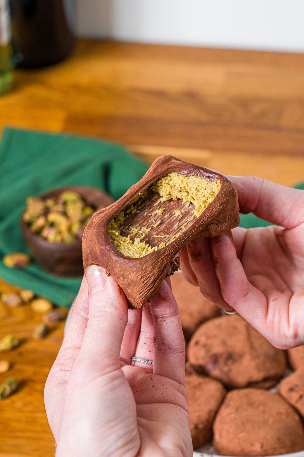 A close up of a hand pulling apart a Dubai chocolate cookie. There is a plate of cookies on a wooden counter with a green cloth napkin and bowl of pistachios blurred in the background.