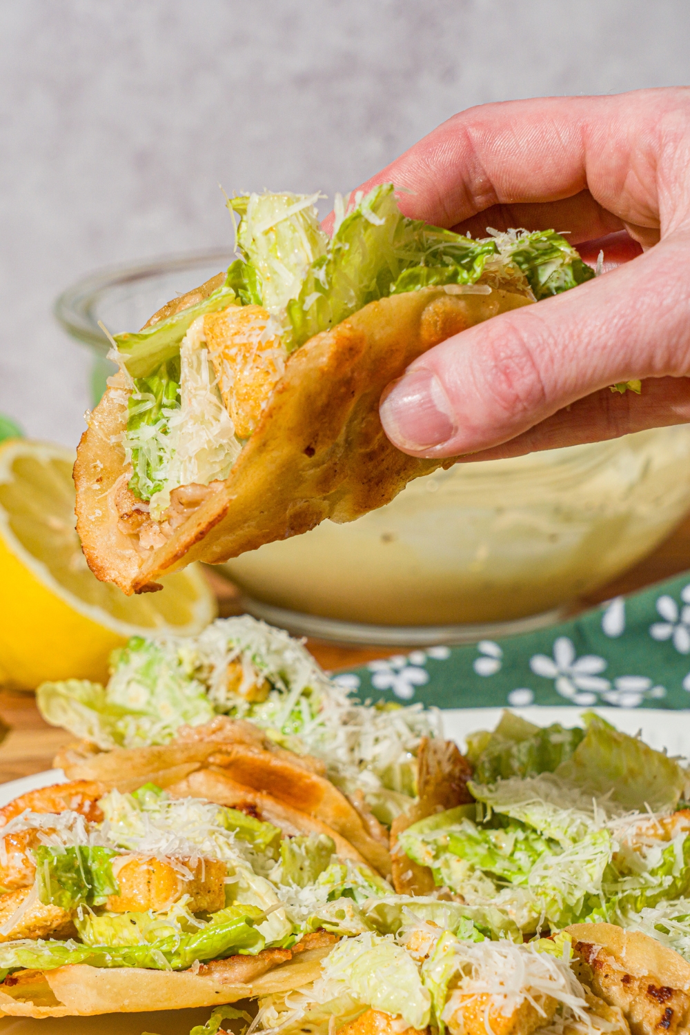 A hand taking a chicken caesar smash taco from a white plate of tacos. The taco is finished with shredded parmesan cheese. The plate is on a wooden counter with a lemon and green floral napkin.