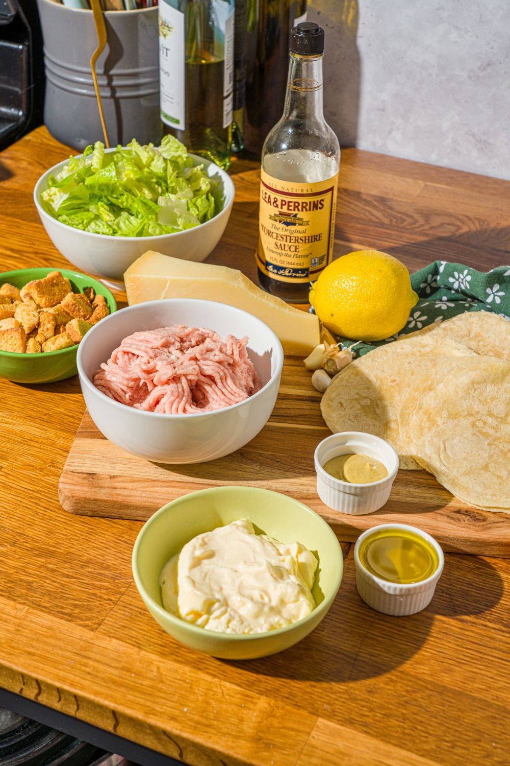 A wooden counter with several bowls containing ingredients to make chicken caesar smash tacos including ground chicken, tortilla wraps, romaine lettuce, lemon, croutons, mayo, olive oil, and mustard.