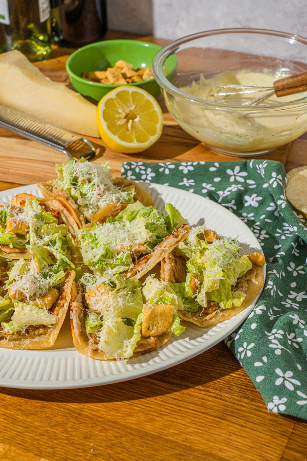 A white plate with several chicken caesar smash tacos finished with shredded parmesan cheese. The plate is on a wooden counter with a green floral napkin, bowl of caesar dressing, and lemon.