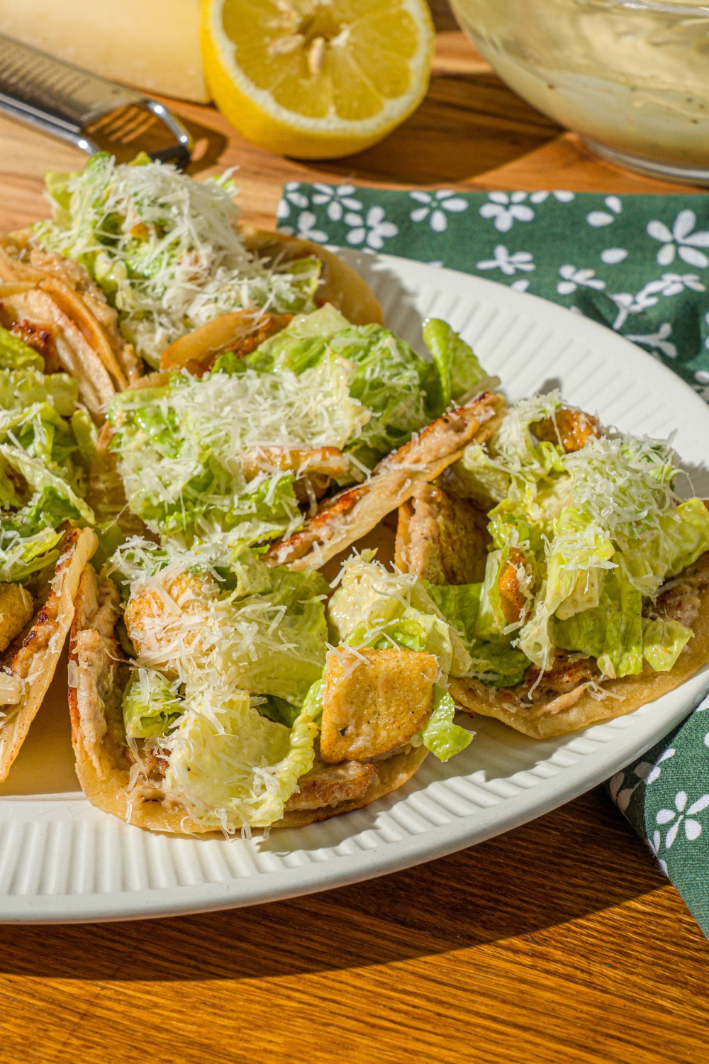 A white plate with several chicken caesar smash tacos finished with shredded parmesan cheese. The plate is on a wooden counter with a green floral napkin, bowl of caesar dressing, and lemon.