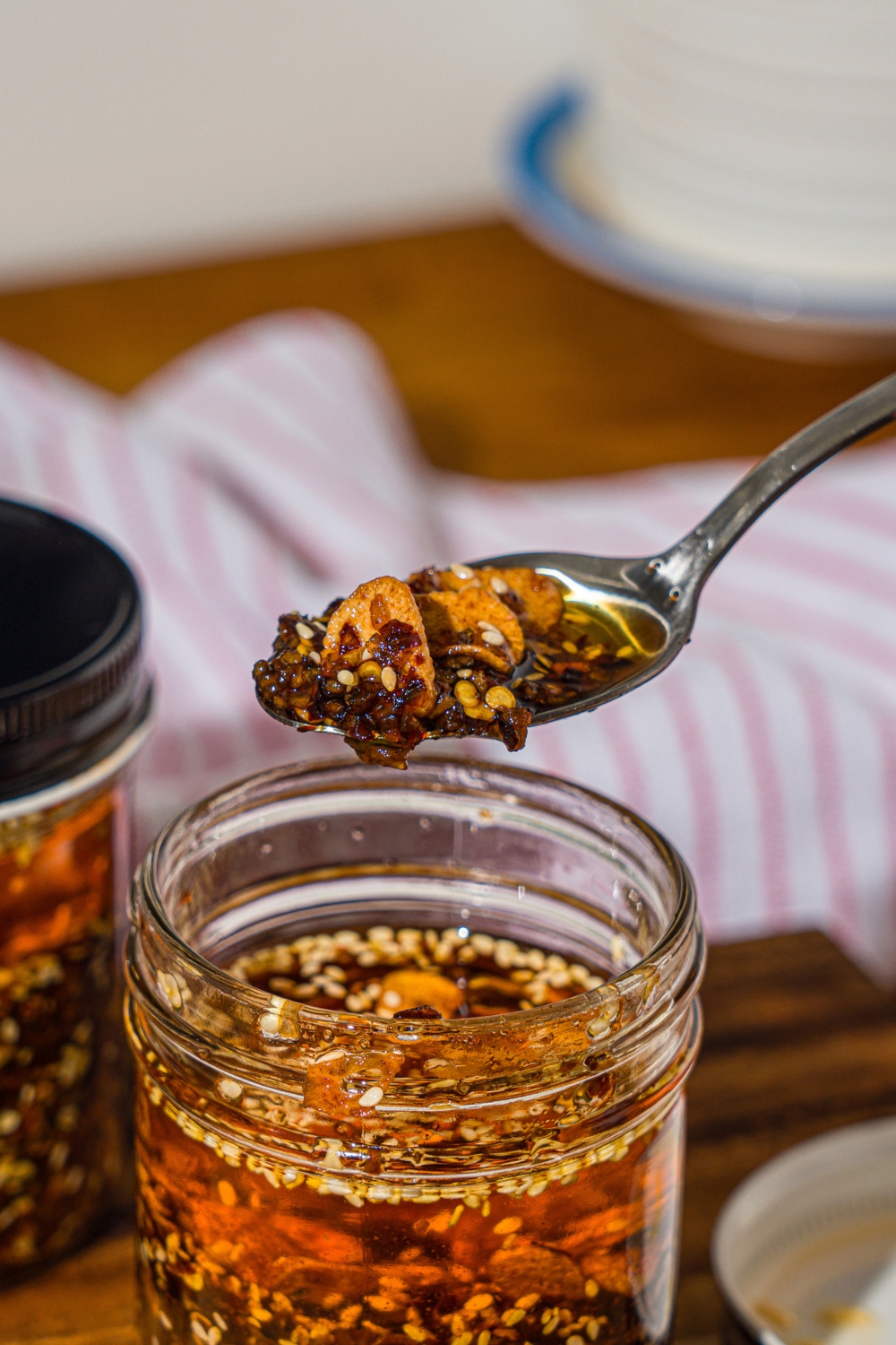 A spoon of chili crisp oil over a jar of chili crisp. The jar is on a wooden board with additional jars of chili crisp oil.