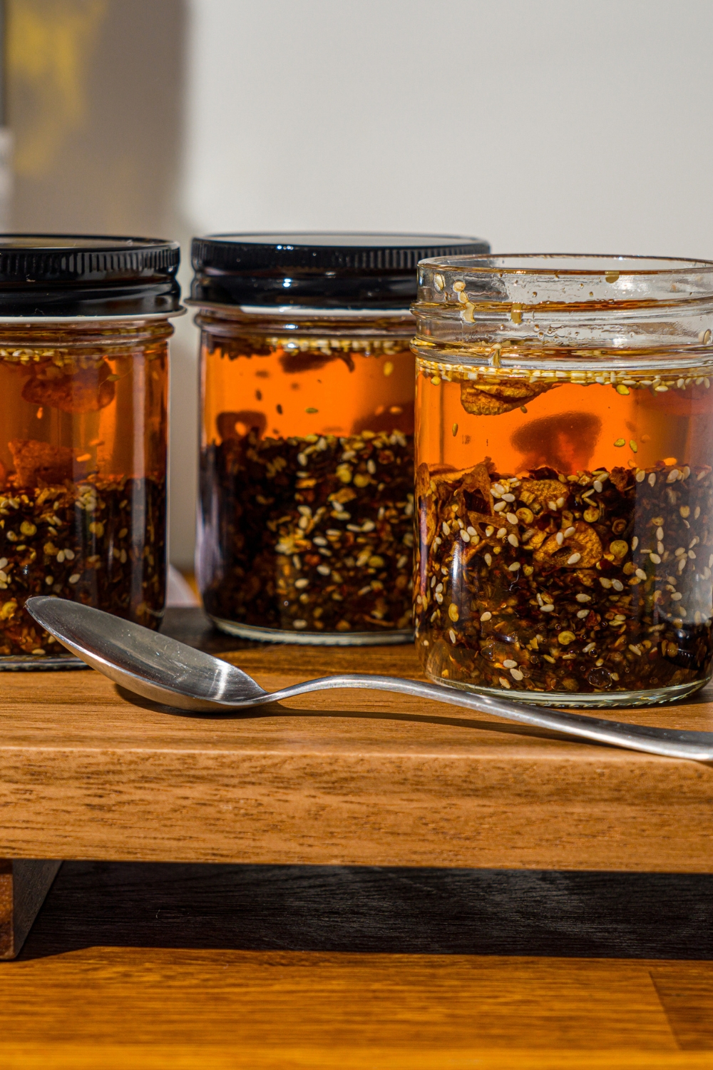 A wooden board with three jars of chili crisp. There is a spoon on the board.