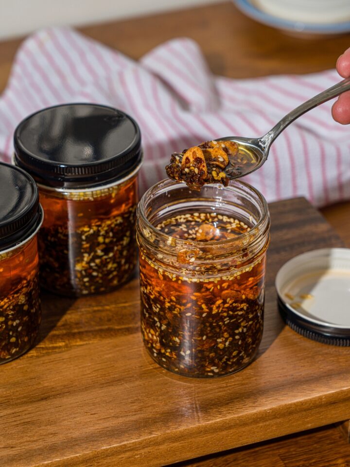 A spoon with chili crisp over a jar of chili crisp oil. The jar is on a wooden board with additional jars of oil.