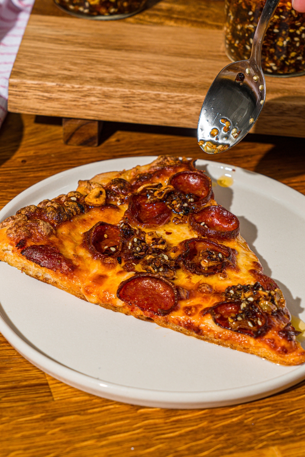 A spoon pouring chili crisp over a white plate with a slice of pepperoni pizza. The plate is on a wooden counter.
