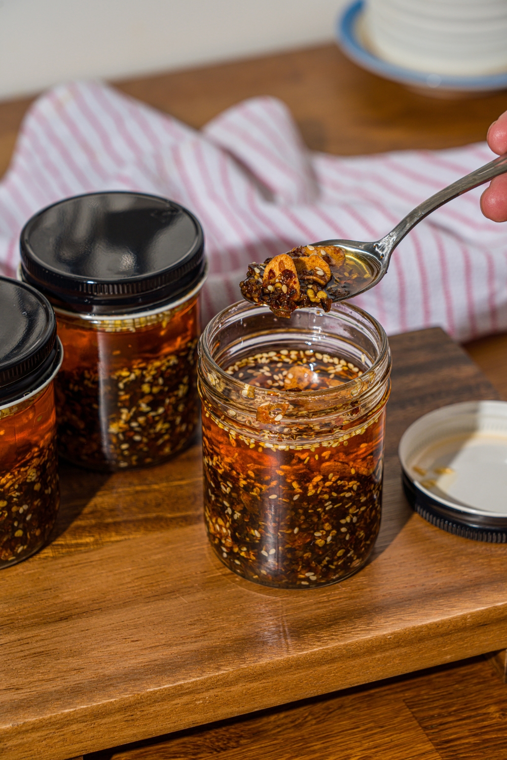 A spoon with chili crisp over a jar of chili crisp oil. The jar is on a wooden board with additional jars of oil.