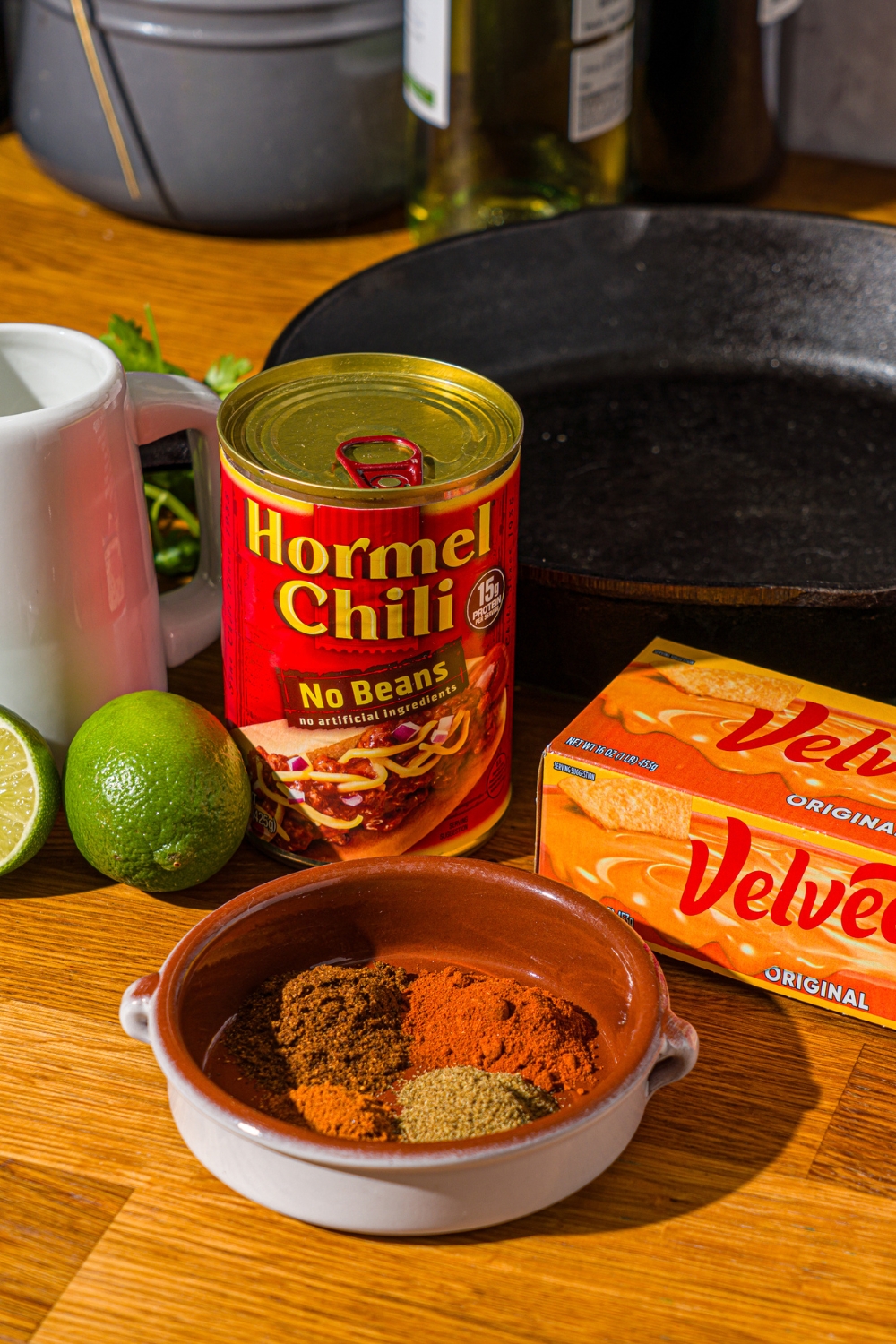 A wooden counter with ingredients to make Chili's skillet queso including Velveeta, Hormel chili, milk, lime, and seasonings.