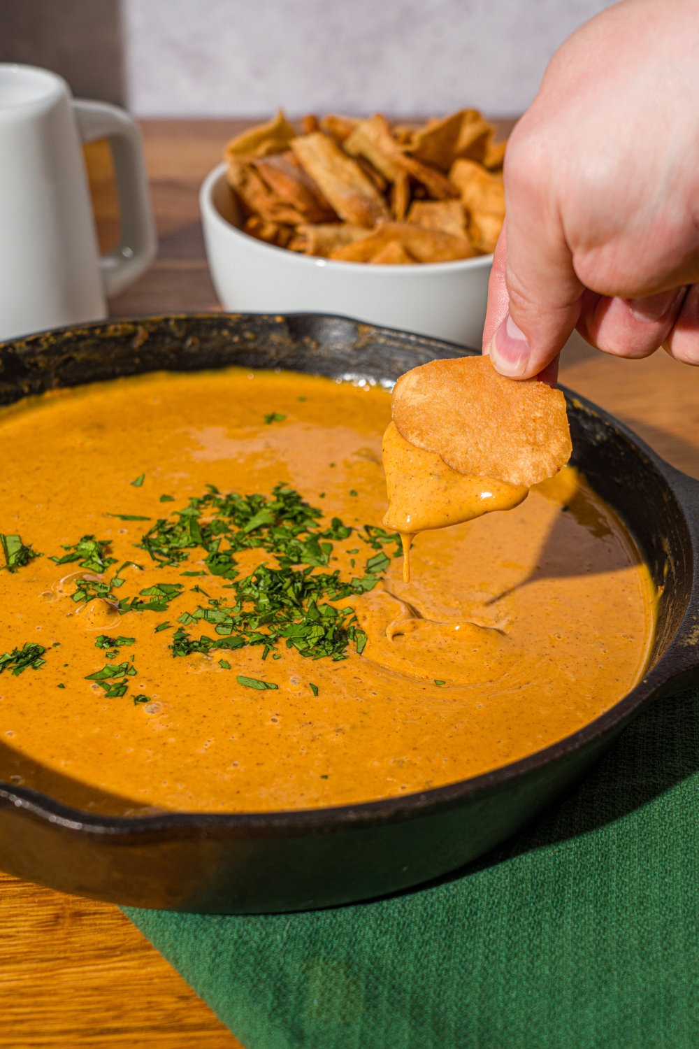 A skillet with Chili's skillet queso garnished with fresh cilantro. A hand is dipping a chip in the queso. The skillet is on a wooden counter with a green cloth napkin and a bowl of chips.