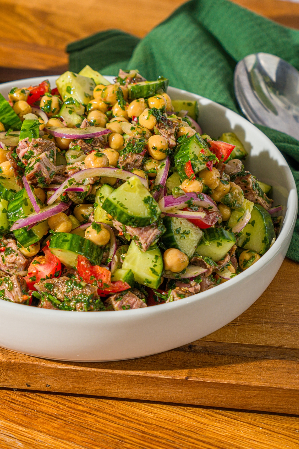 A white bowl with chimichurri steak salad tossed with beans, cucumbers, tomatoes, and red onion in a chimichurri dressing. The bowl is on a wooden counter with a green cloth napkin.