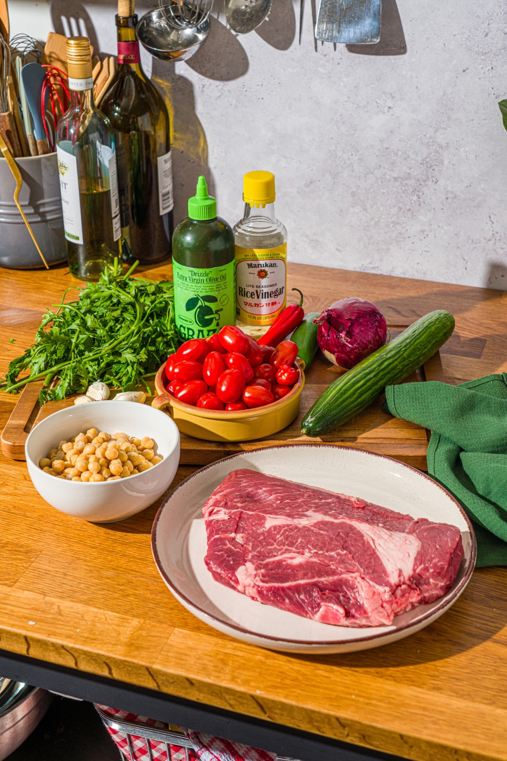 A wooden counter with ingredients to make chimichurri steak salad including steak, chick peas, tomatoes, cucumbers, fresh parsley, red onion, and oil.