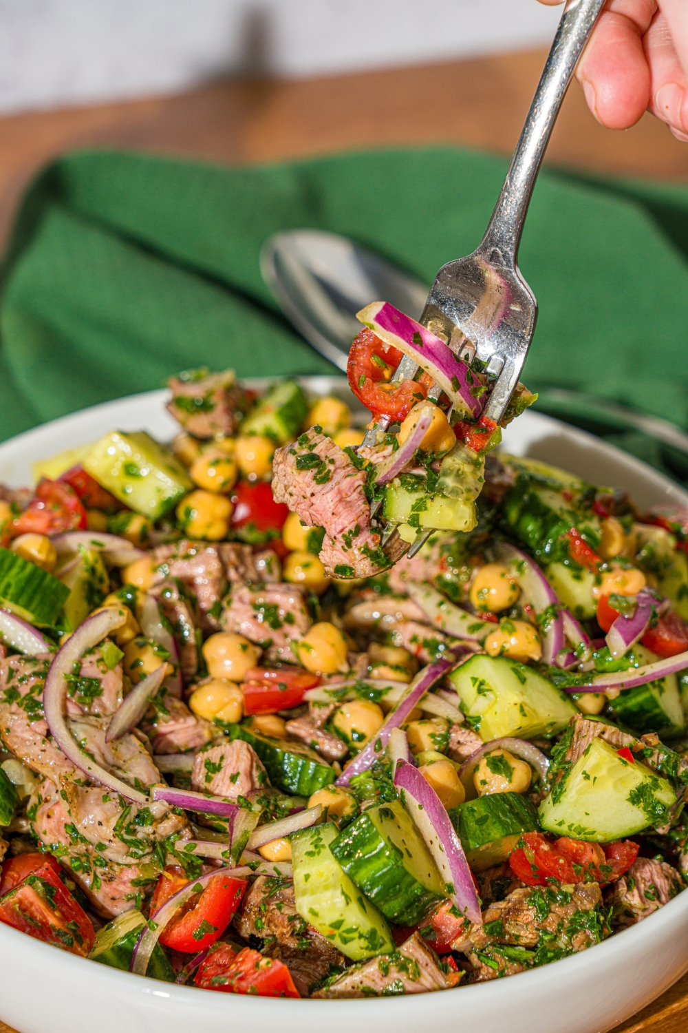 A white bowl with chimichurri steak salad tossed with beans, cucumbers, tomatoes, and red onion in a chimichurri dressing. There is a fork taking a bite from the bowl. The bowl is on a wooden counter with a green cloth napkin.
