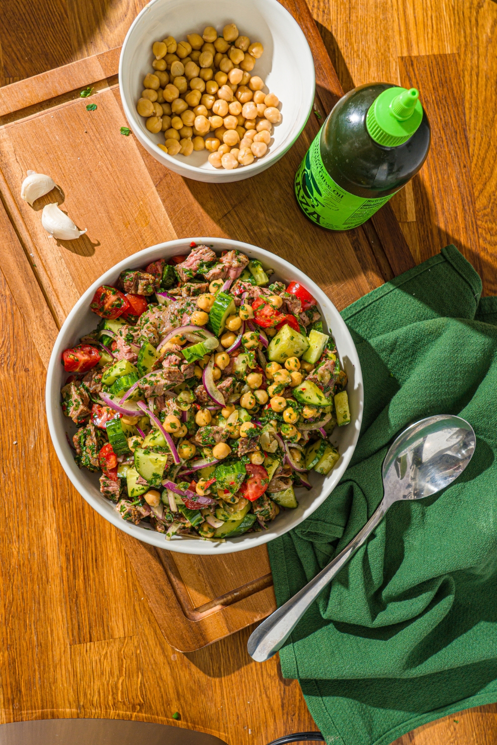 A white bowl with chimichurri steak salad tossed with beans, cucumbers, tomatoes, and red onion in a chimichurri dressing. The bowl is on a wooden counter with a green cloth napkin and a bowl of chick peas.