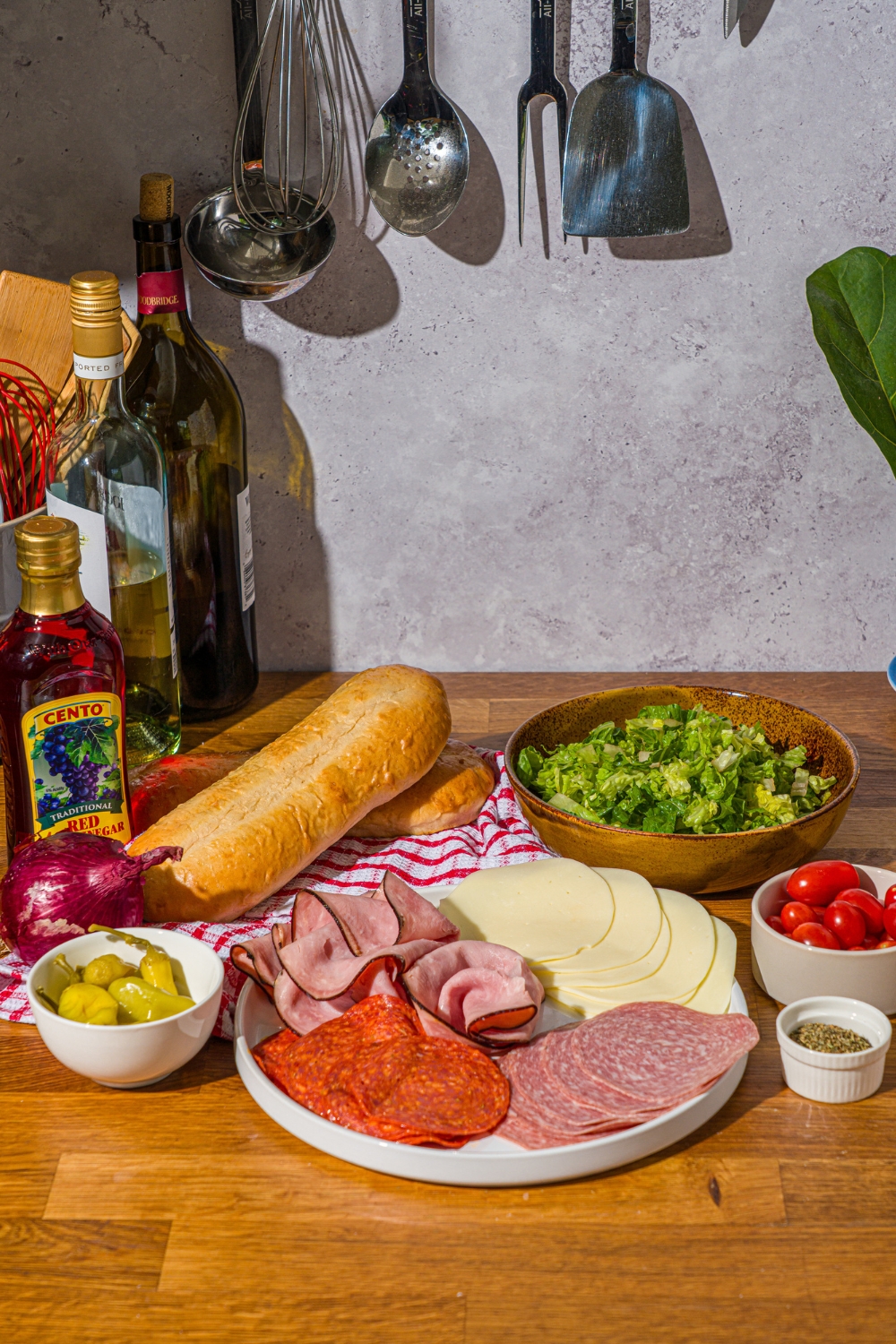 A wooden counter with ingredients to make a chopped Italian sandwich including salami, provolone, pepperoni, deli ham, chopped lettuce, tomato, red onion, pepperocini peppers, a hoagie roll, mayo, and seasonings.