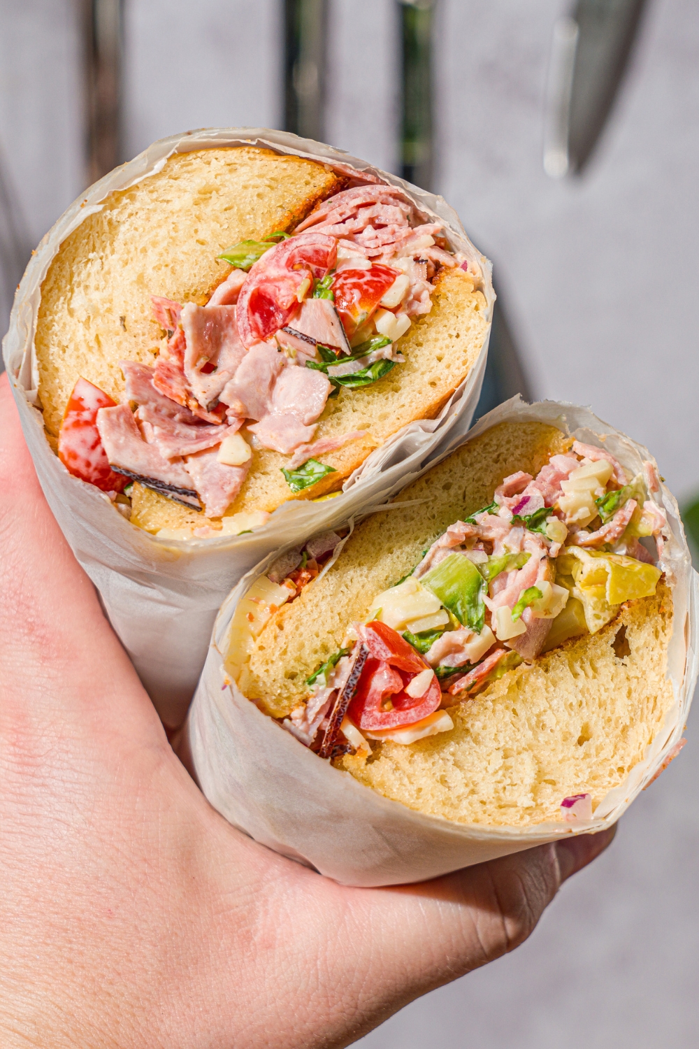 A hand holding a chopped Italian sandwich wrapped in parchment paper and sliced in half.